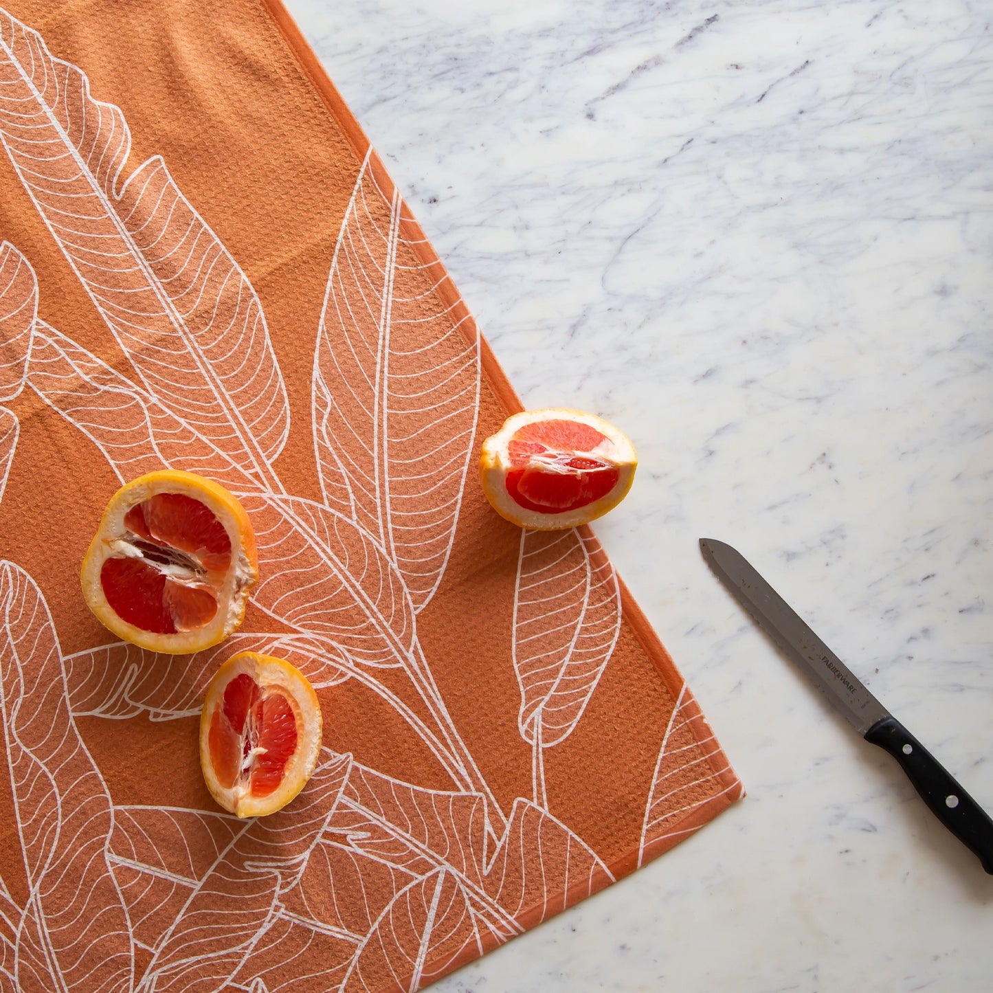 On a marble background is an orange kitchen towel with a white banana leaf design.