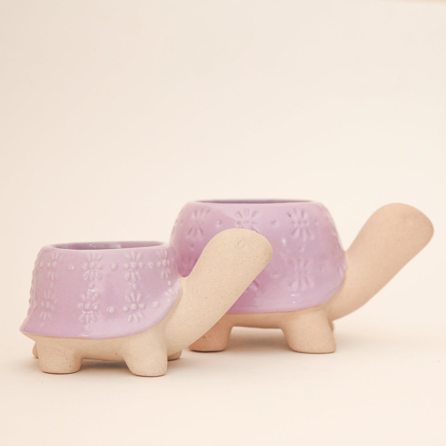 On a light peach background is two different sized ceramic planters in the shape of turtles with a subtle flower pattern on the shells.