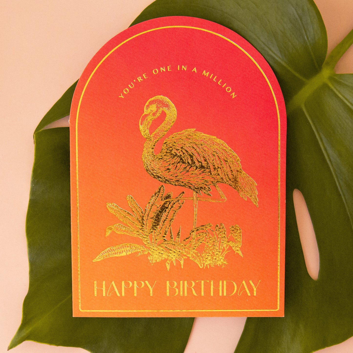 An orange gradient arched shaped card with gold foiled illustrations of a flamingo and text that reads, "You're One In A Million Happy Birthday".