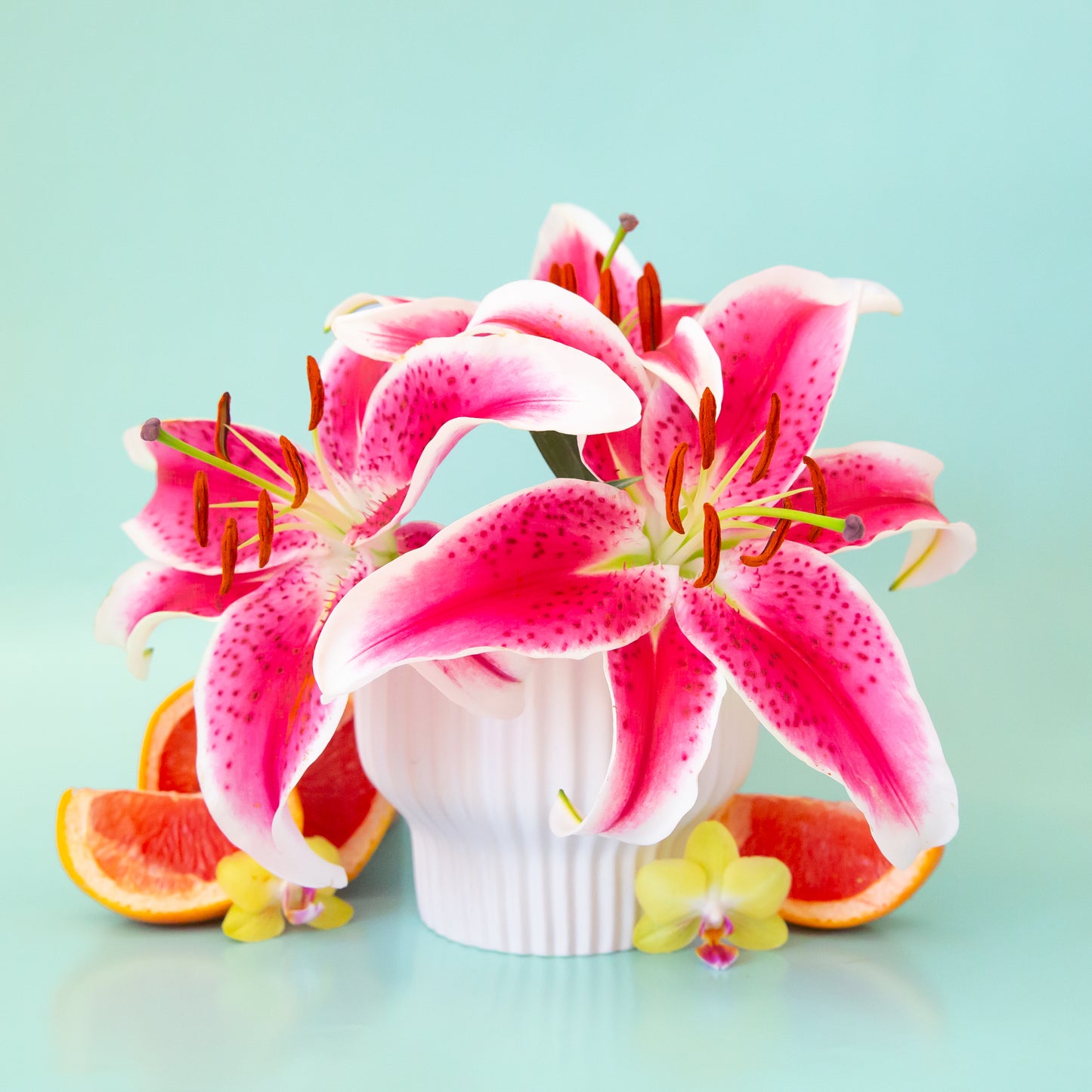 On a blue background is a white ribbed pedestal planter filled with lily flowers not included with purchase.
