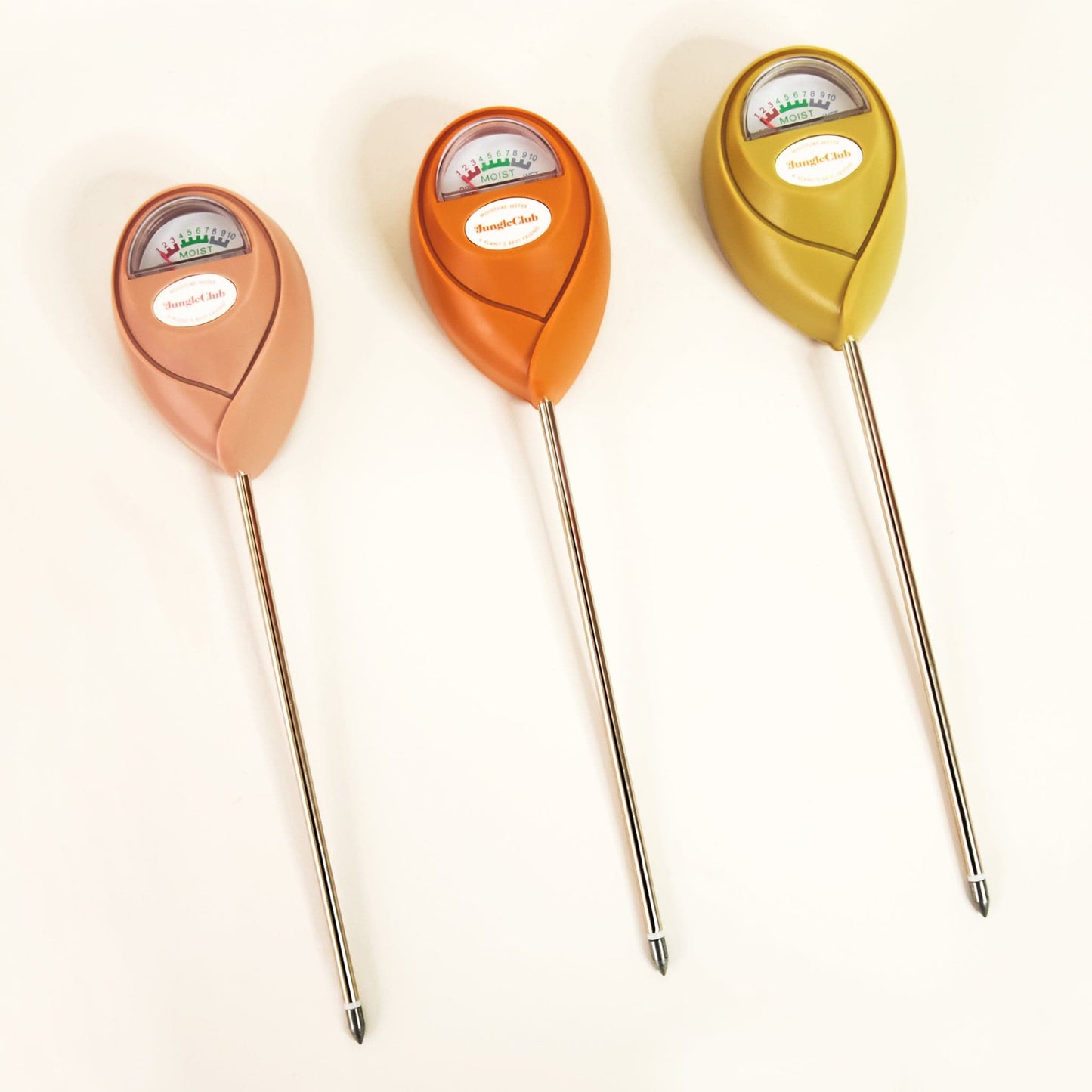 On a cream background is three different colored moisture meters, one pink, one orange and one tropical green. They feature a long pointed metal bottom that sticks into the plant's soil and a meter at the top with a moisture gauge and a white oval in the center that reads, "Jungle Club".
