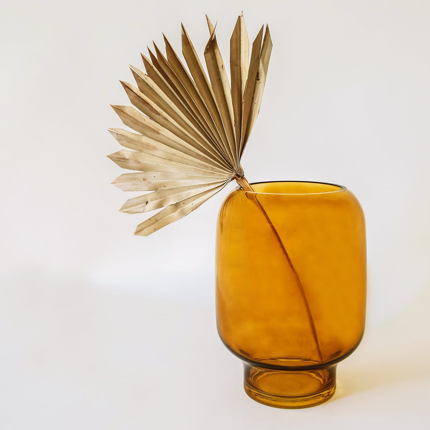On a white background is an amber glass vase with a single dried palm leaf displayed inside.