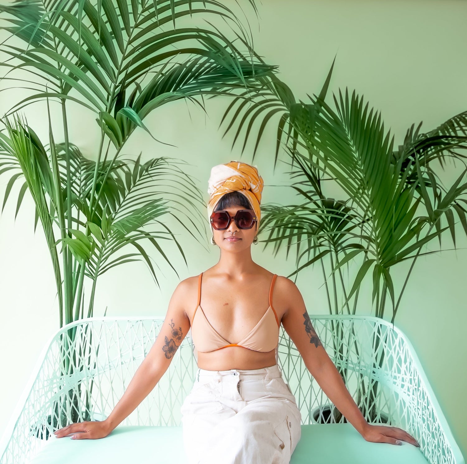 A woman sits in the middle of frame sitting on a white bench.  She has her arms outstretched on a white bench she is seated on.  Behind her are two palm trees.  She has large sunglasses on and her hair wrapped in an orange towel on top of her head.