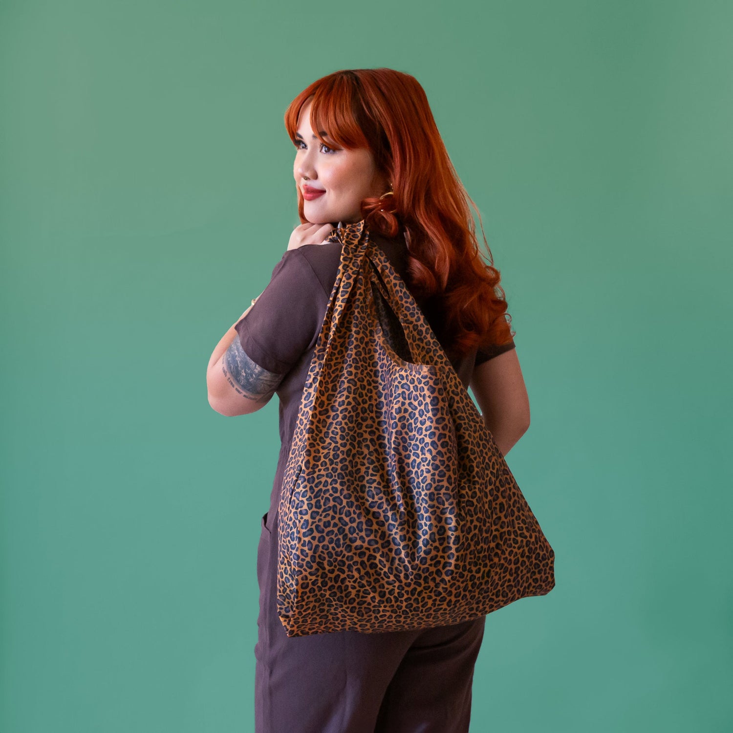 A woman with beautiful red hair stands with her back to the camera.  She has a leopard print reusable bag slung over her left shoulder.  The background of the image is a beautiful jade green color.