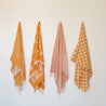On an ivory background is four beach towels hung up. From left to right, the towels are the Banana Leaf Beach Towel, Retro Flower Beach Towel, Bird of Paradise Beach Towel and the Checker Beach Towel.