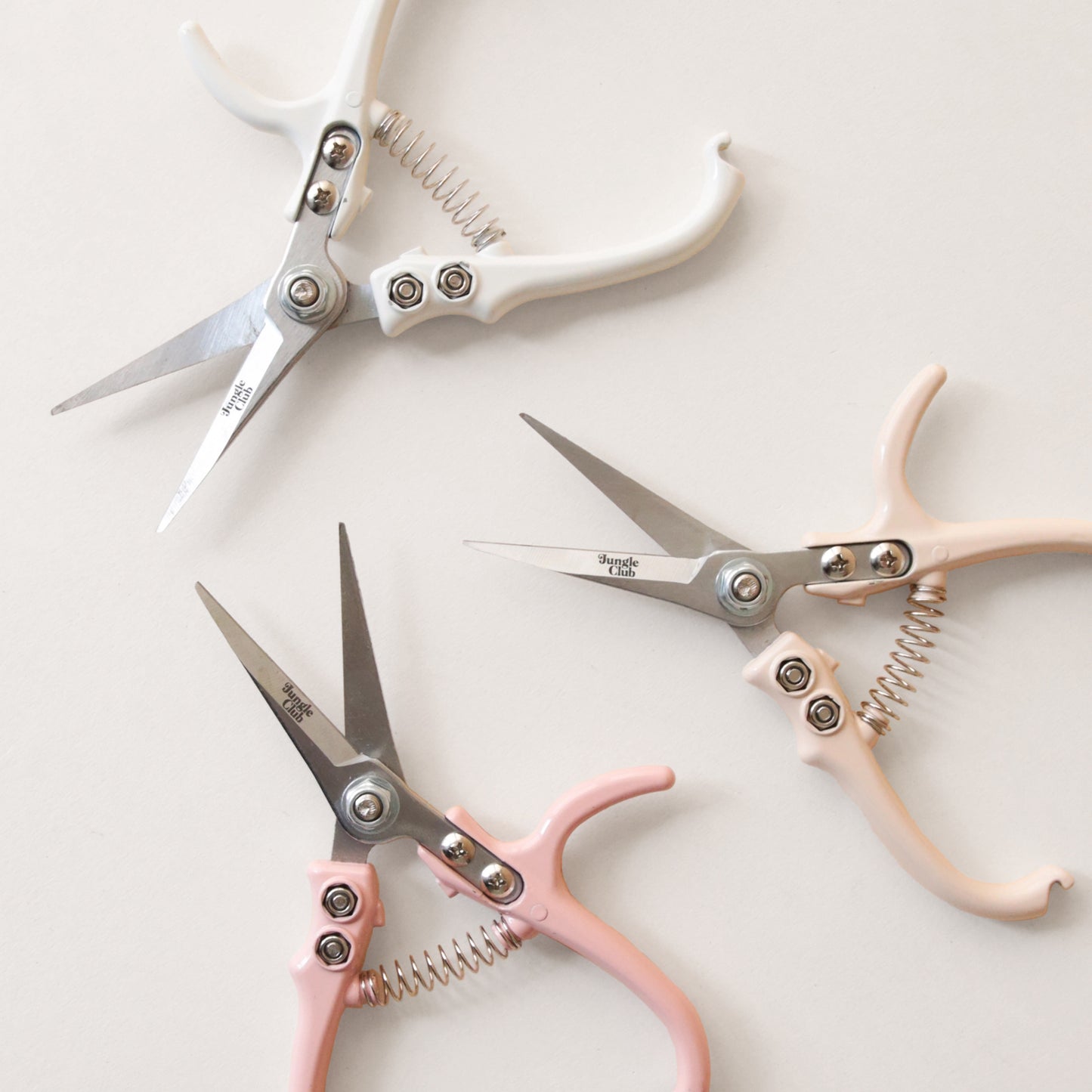Pruning shears with a narrow tip, and light cream colored handle and a black clasp to connect when shears are not in use. There is small black text on the side of the shears that read, "Jungle Club" photographed here with the other colors available, pink and white.