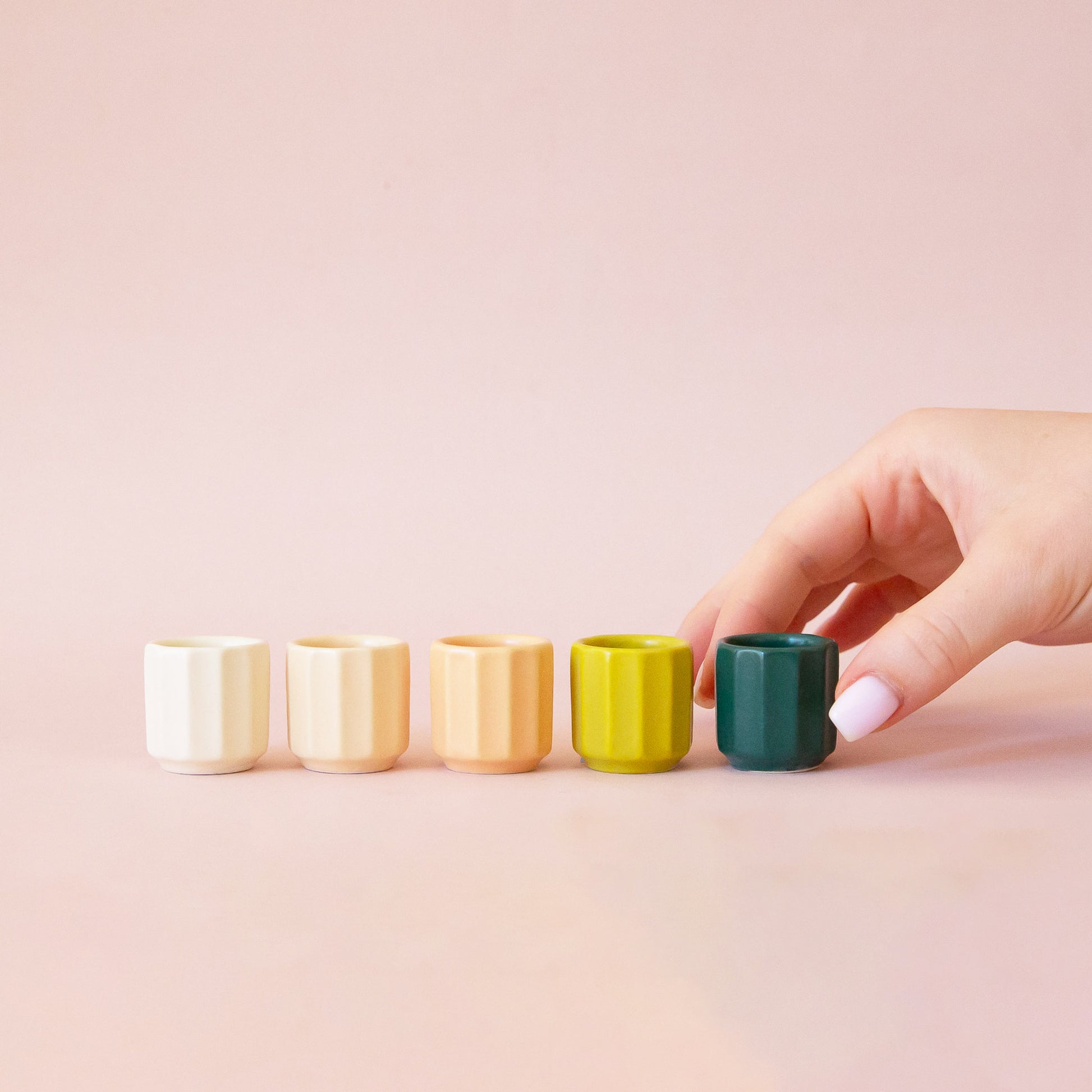 A hand comes into frame on the right hand side of the image.  The hand reaches for a miniature aquamarine colored pot.  The pot is last in a row of 5 tiny pots.  They go in order from white to ivory to peach to chartreuse to the final aquamarine color.  The background of the image is a soft pink color.