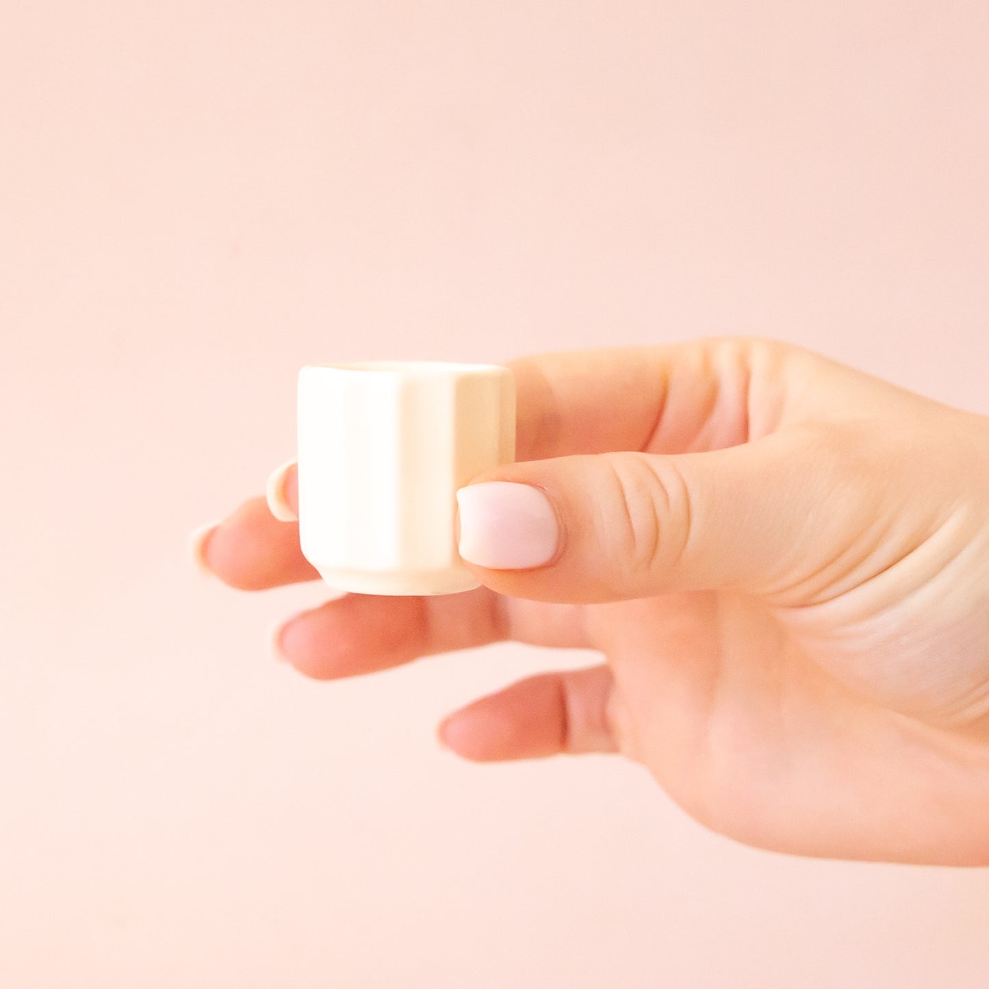 A hand comes into frame on the right hand side of image with soft pink nails.  The hand is holding an itty bitty pot about the size of thimble that is a white color.  The background is all  a soft white color.