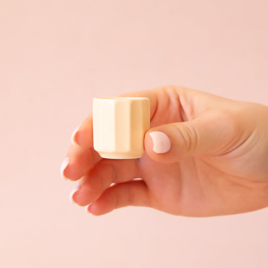 A hand comes into frame on the right hand side of image with soft pink nails.  The hand is holding an itty bitty pot about the size of thimble that is a soft tan color.  The background is all  a soft white color.