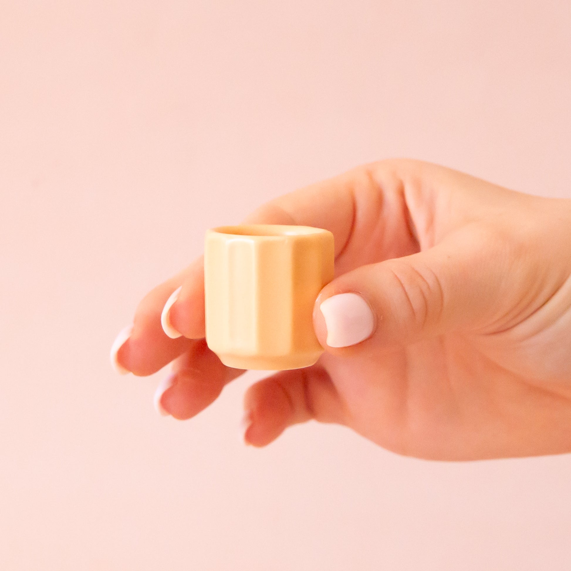 A hand comes into frame on the right hand side of image with soft pink nails.  The hand is holding an itty bitty pot about the size of thimble that is a soft peach color.  The background is all  a soft white color.