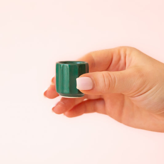 A hand comes into frame on the right hand side of image with soft pink nails.  The hand is holding an itty bitty pot about the size of thimble that is a dark blue green color.  The background is all  a soft white color.