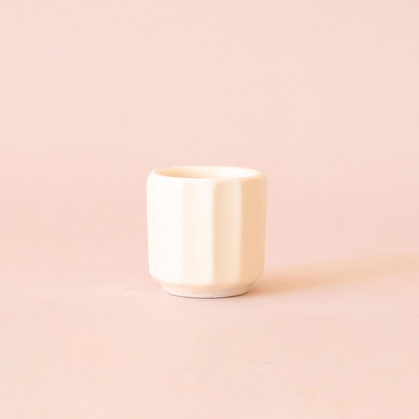 An itty bitty pot about the size of thimble that is a white color sits in the middle of a page.  The background is a soft pink color.