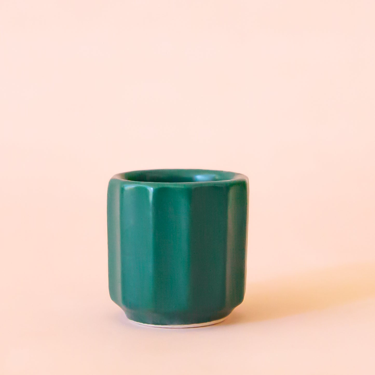  An itty bitty pot about the size of thimble that is a dark blue green color sits in the middle of the image.  The pot hat little ridges running around it.  The background of the image is a soft white color.