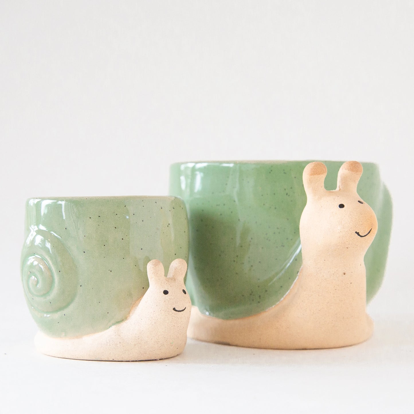 On a light grey background is two ceramic planters in the shape of a snail with smiling faces and a swirly green "shell".