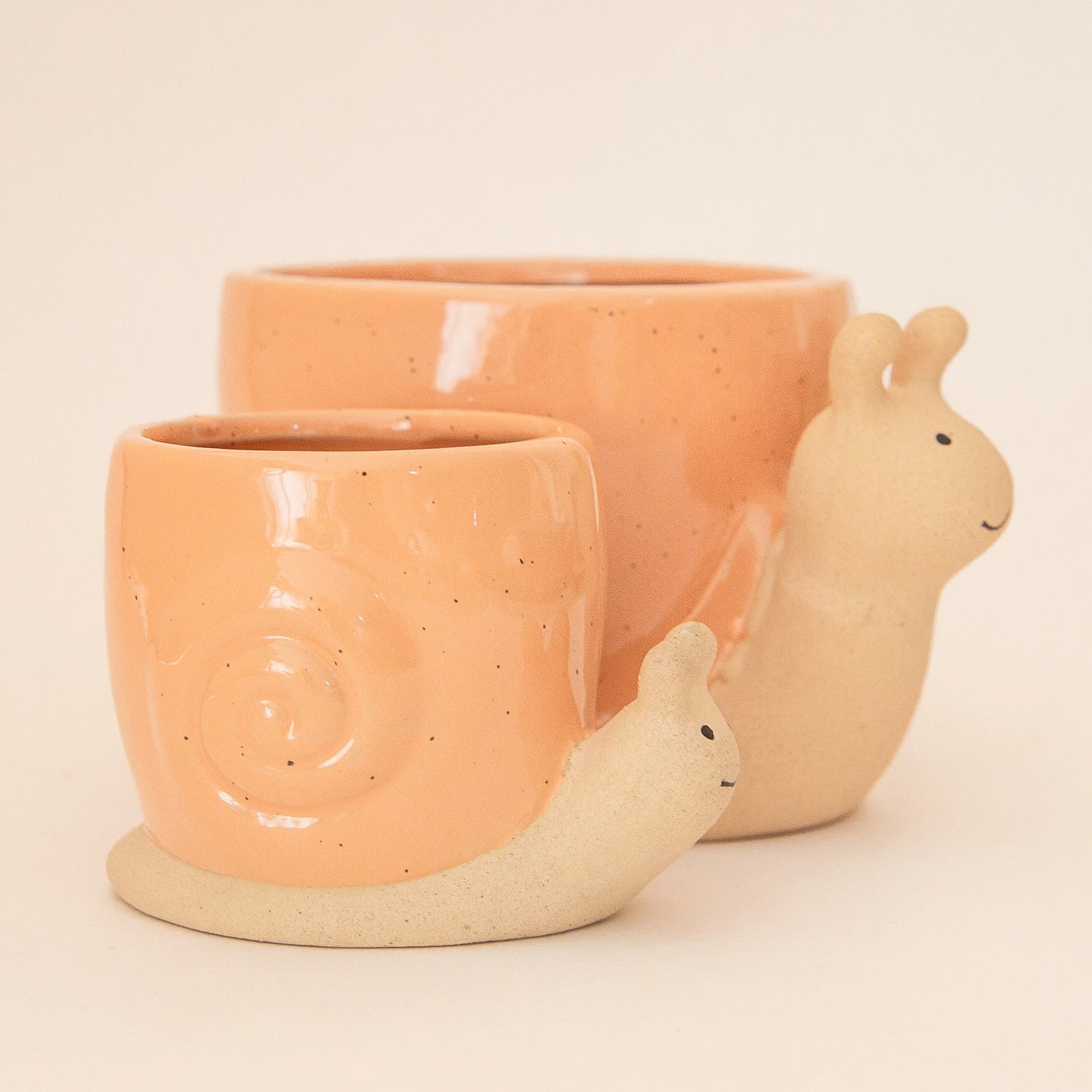 On a light peach background is two ceramic planters in the shape of a snail with a swirly light orange "shell".
