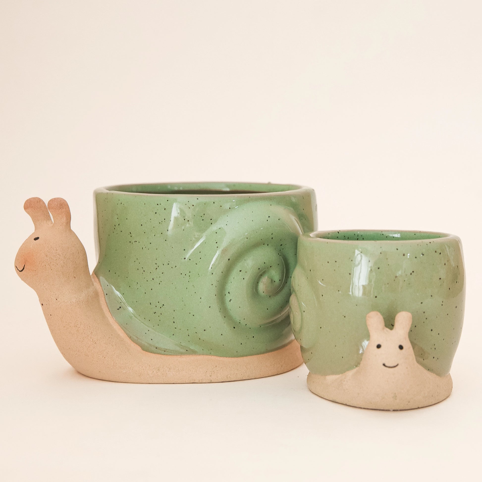On a tan background is two ceramic planters in the shape of a snail with smiling faces and a swirly green "shell".