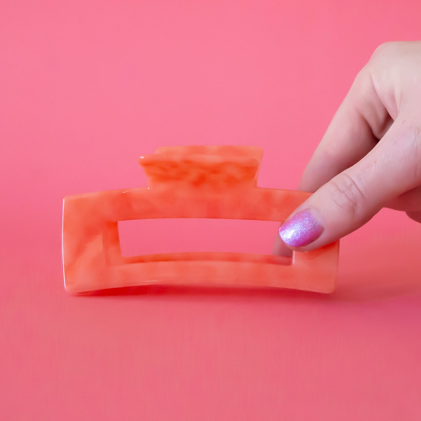 A beautiful coral colored hair claw sits in the middle of page.  A hand reaches into frame from the right hand side holding onto the hair claw.  The thumb nail is painted with glitter.  The background is a watermelon color.