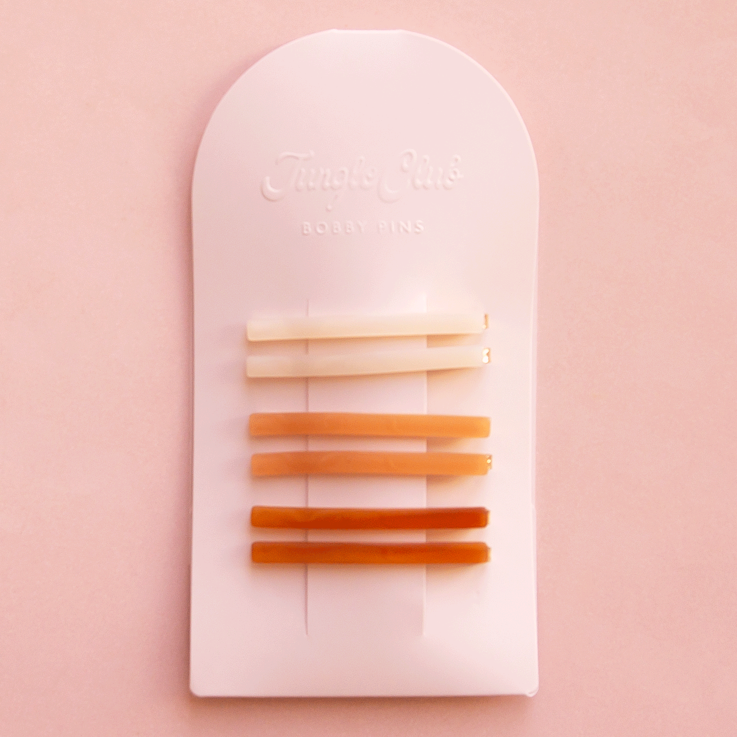 On a pink background is an arched packaging with embossed text at the top that reads, "Jungle Club Bobby Pins" along with six thin hair clips in three colors from an ivory shade to orange to a honey rust color.