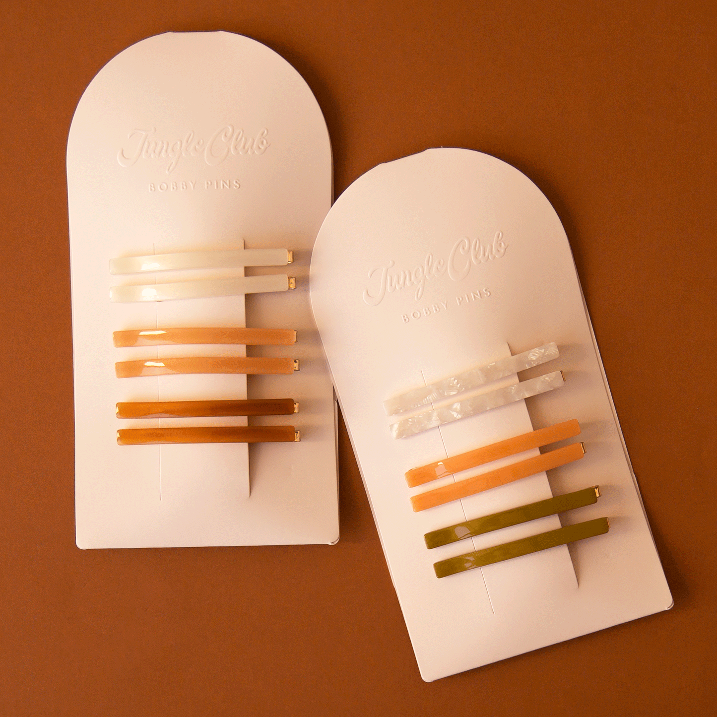 On a burnt orange background is both of the available color ways of the acetate bobby pin sets on arched packaging.