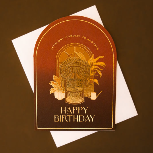 An arched card with orange and brown hues with gold foiled text that reads, 'From One Goddess To Another Happy Birthday' and a design of a peacock chair with plants behind it. 