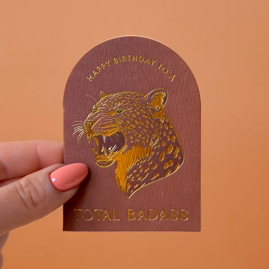 An arched brown card with gold foiled text that reads, 'Happy Birthday to a Total Badass' and a leopard design. 