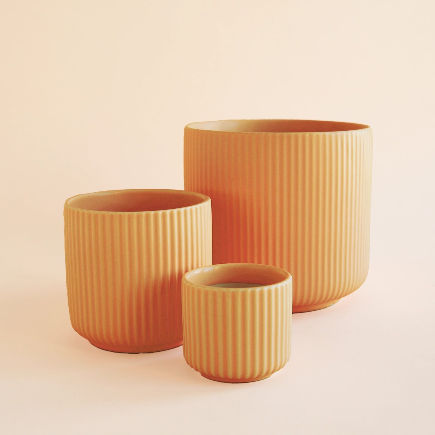 On an ivory background is three different sized orange ceramic planters with a fluted detail and a rounded base. 