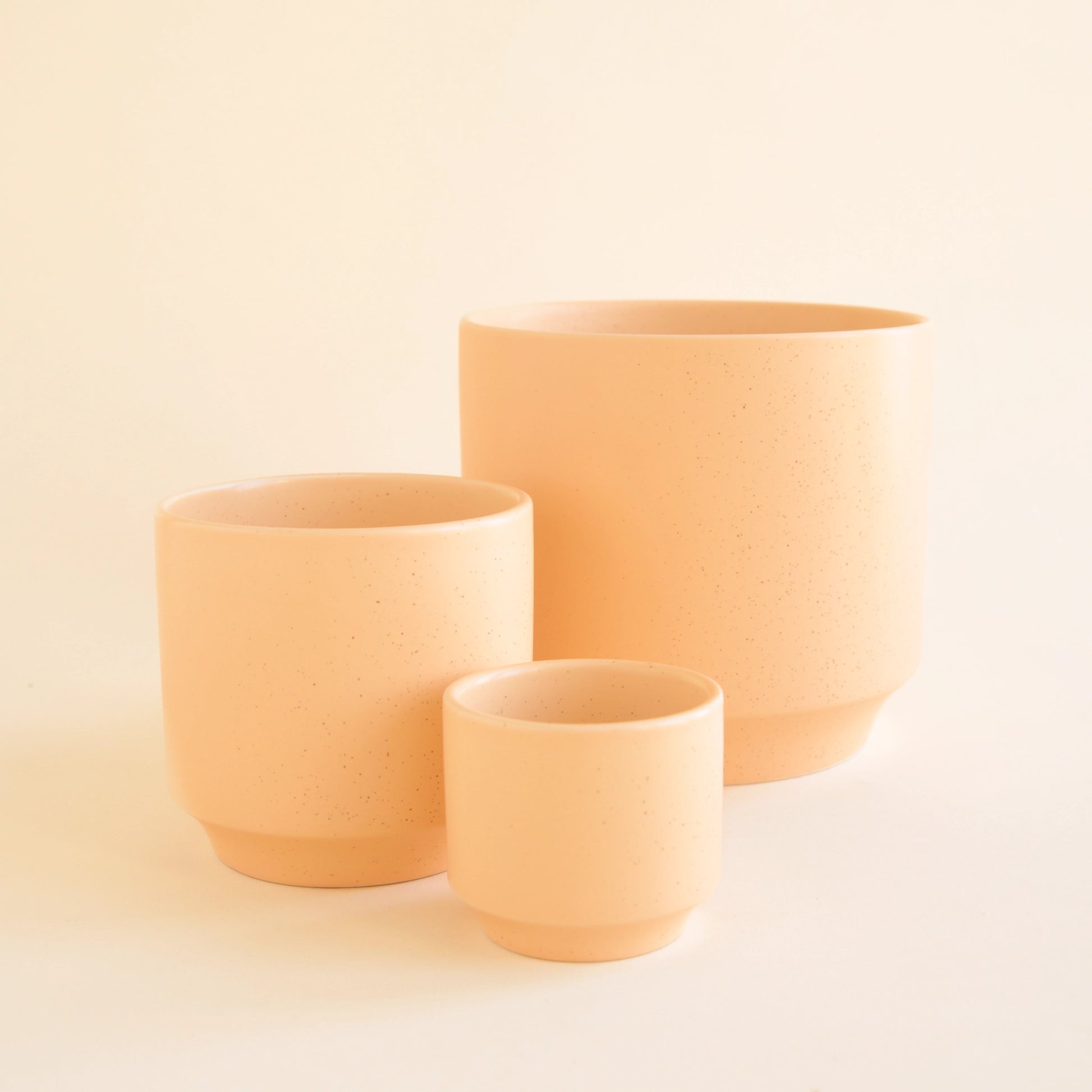 On an ivory background is three different sized ceramic planters in a salmon, pinkish orange shade with a speckle detail and a tapered base. 