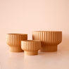 On a peachy background is a burnt orange colored ribbed pedestal planter in three different sizes.
