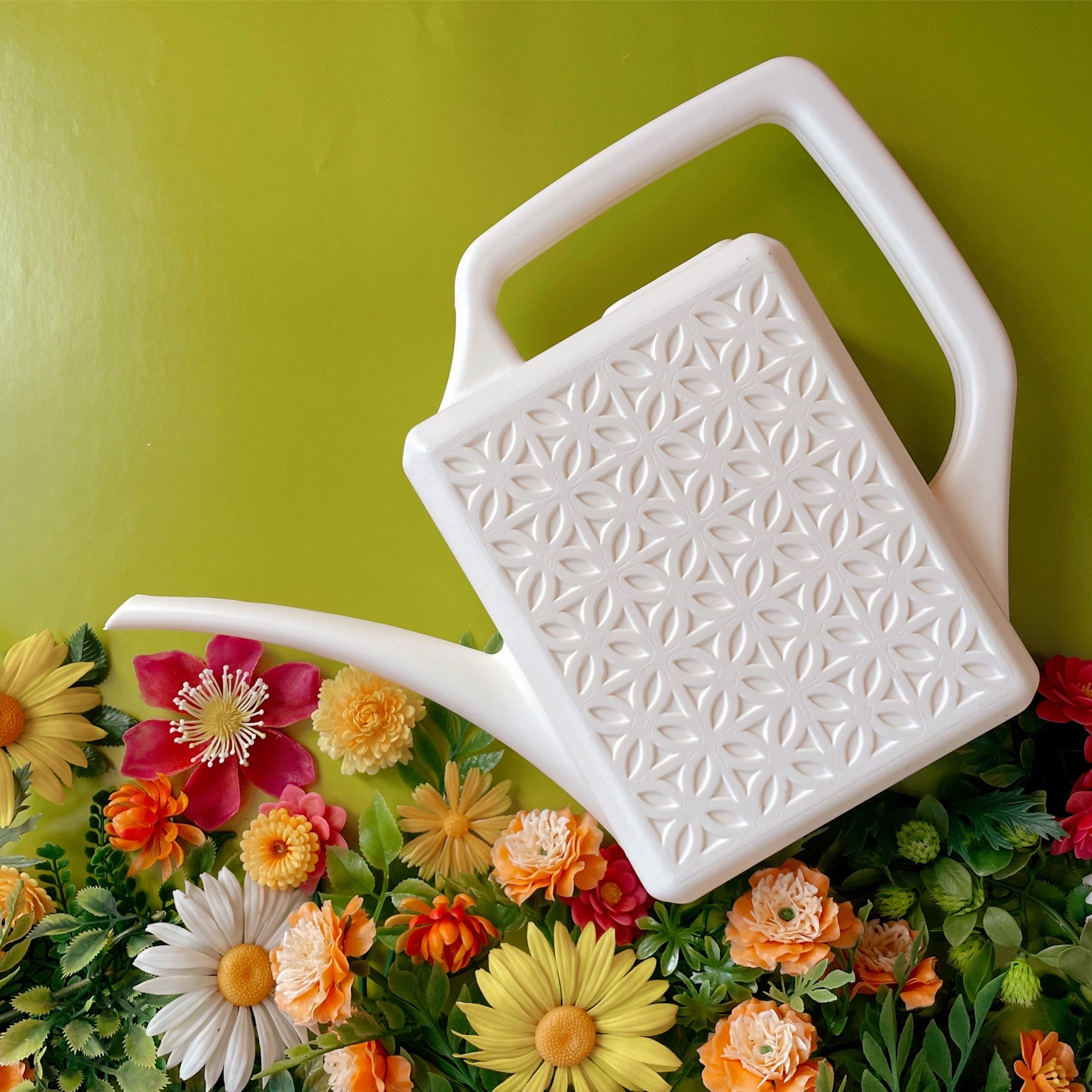A white watering can with a breeze block design on the sides of the watering can. In this photo the watering can is above a bed of flowers. 