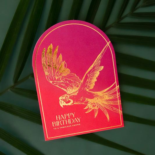 A hot pink and orange ombre colored arched shaped card with gold foiled text that reads, "Happy Birthday To A Fabulous Friend" along with a gold foiled parrot illustration. 