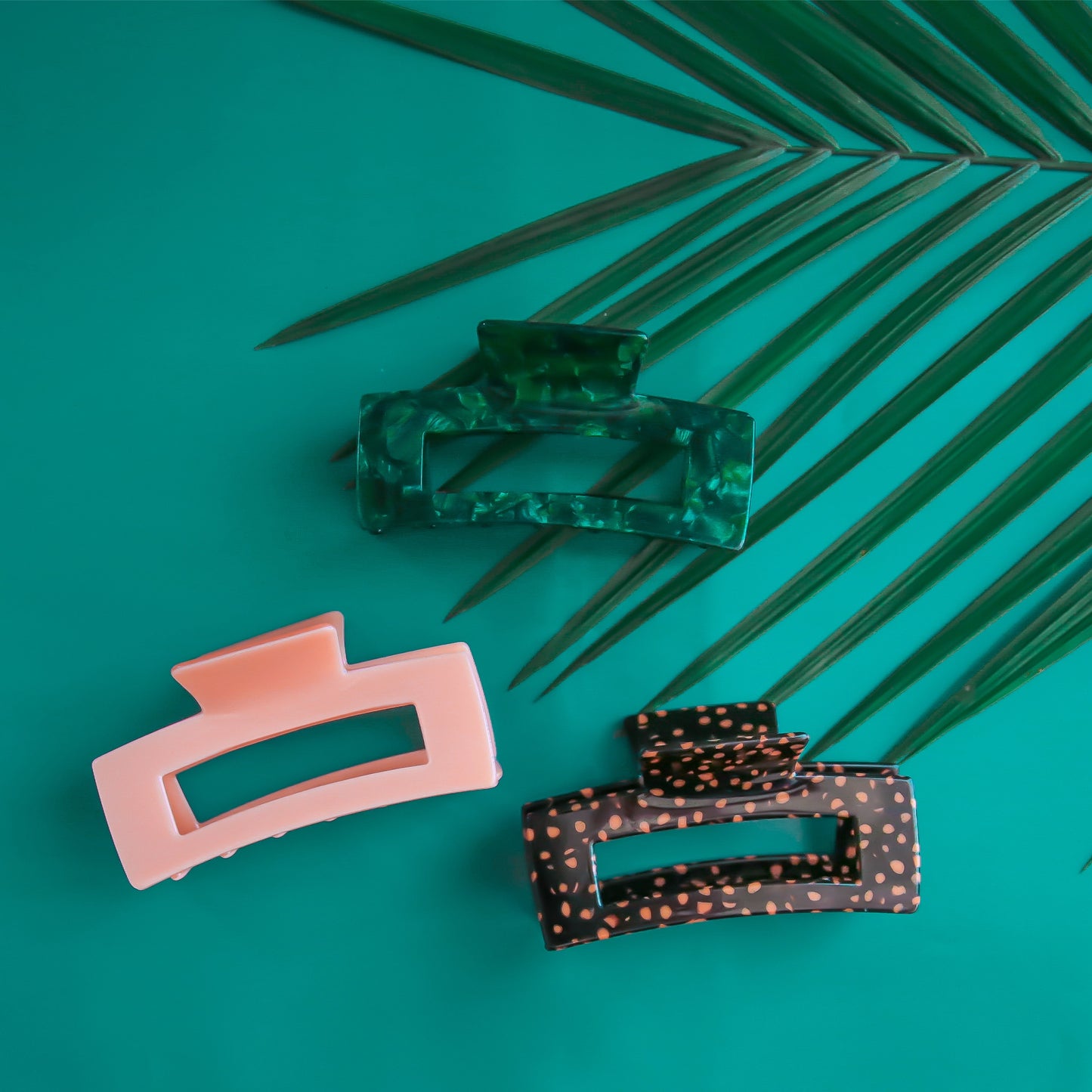 On a turquoise background is the three different colors available for the Paradise Hair Claw. There is an orange and dark brown leopard print, a dark green shade, and a peachy pink option.