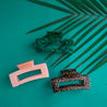On a turquoise background is the three different colors available for the Paradise Hair Claw. There is an orange and dark brown leopard print, a dark green shade, and a peachy pink option.
