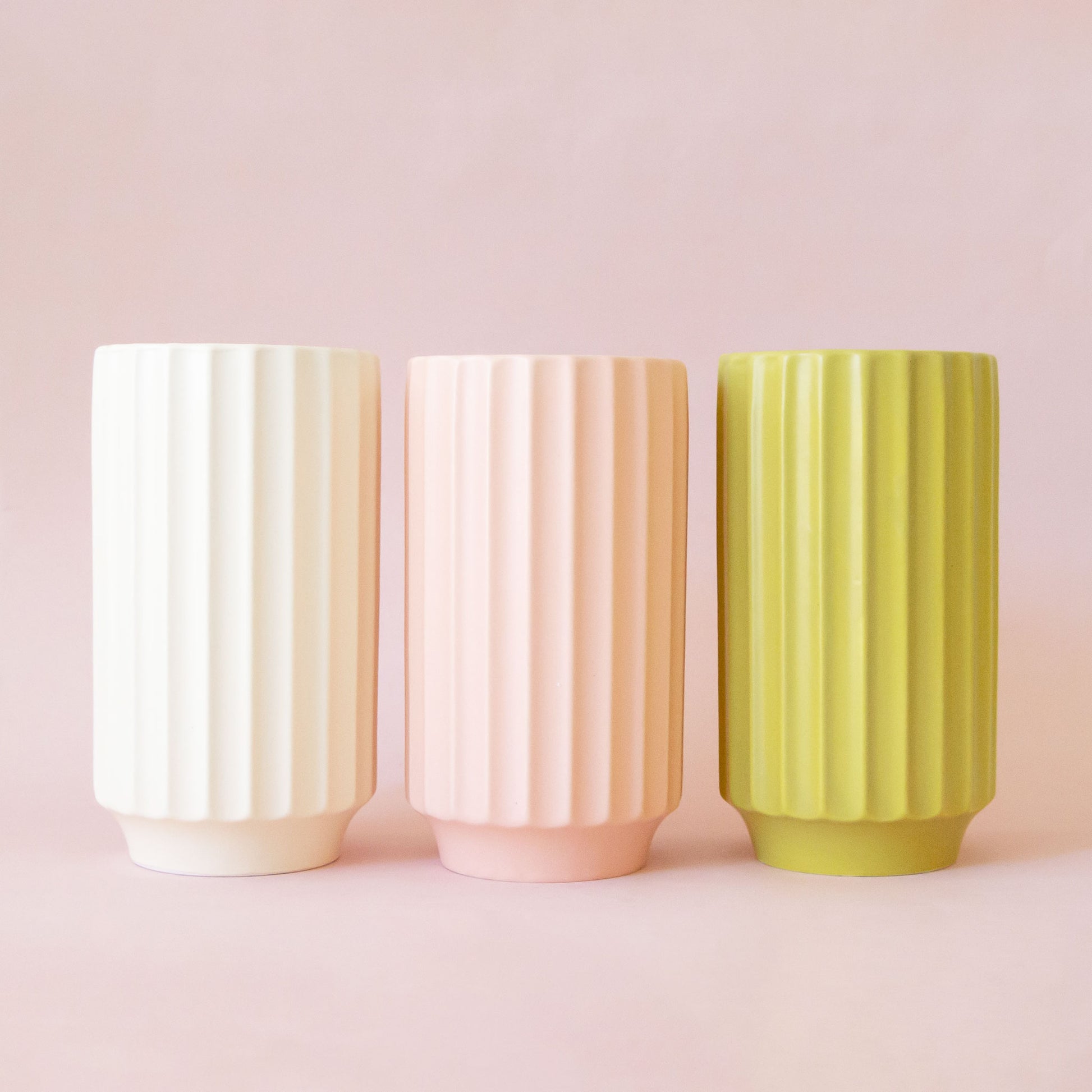 Three beautiful different colored fluted cylindrical vase made our of a ceramic material. Each color sold separately. From left to right the colors are, Vintage White, Petal Pink and Palm. 