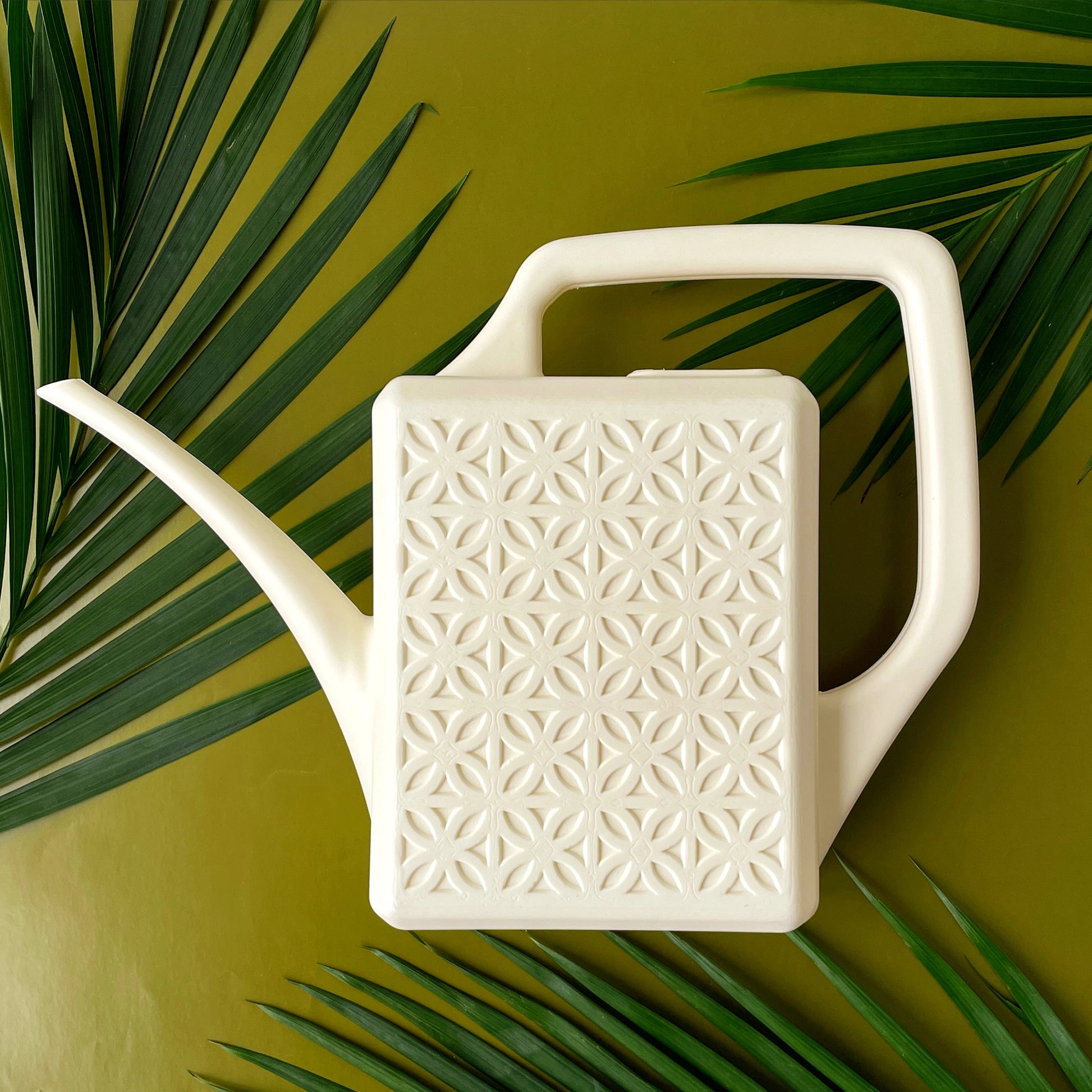 An ivory plastic watering can with a narrow spout and square handle and a rectangle breeze block design on the sides. The watering can is photographed on a lime green ground with palm fronds spilling into the image.
