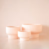 On a peachy background is a light pink ribbed pedestal planter in three different sizes.