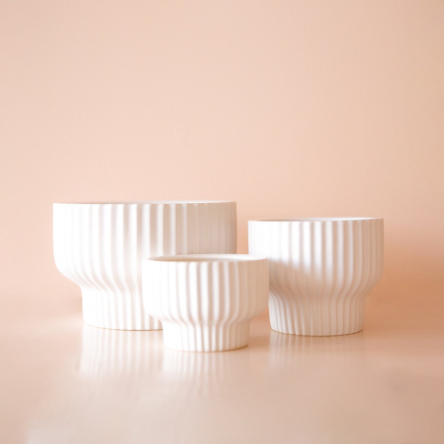 On a peachy background is a white ribbed pedestal planter in three different sizes.