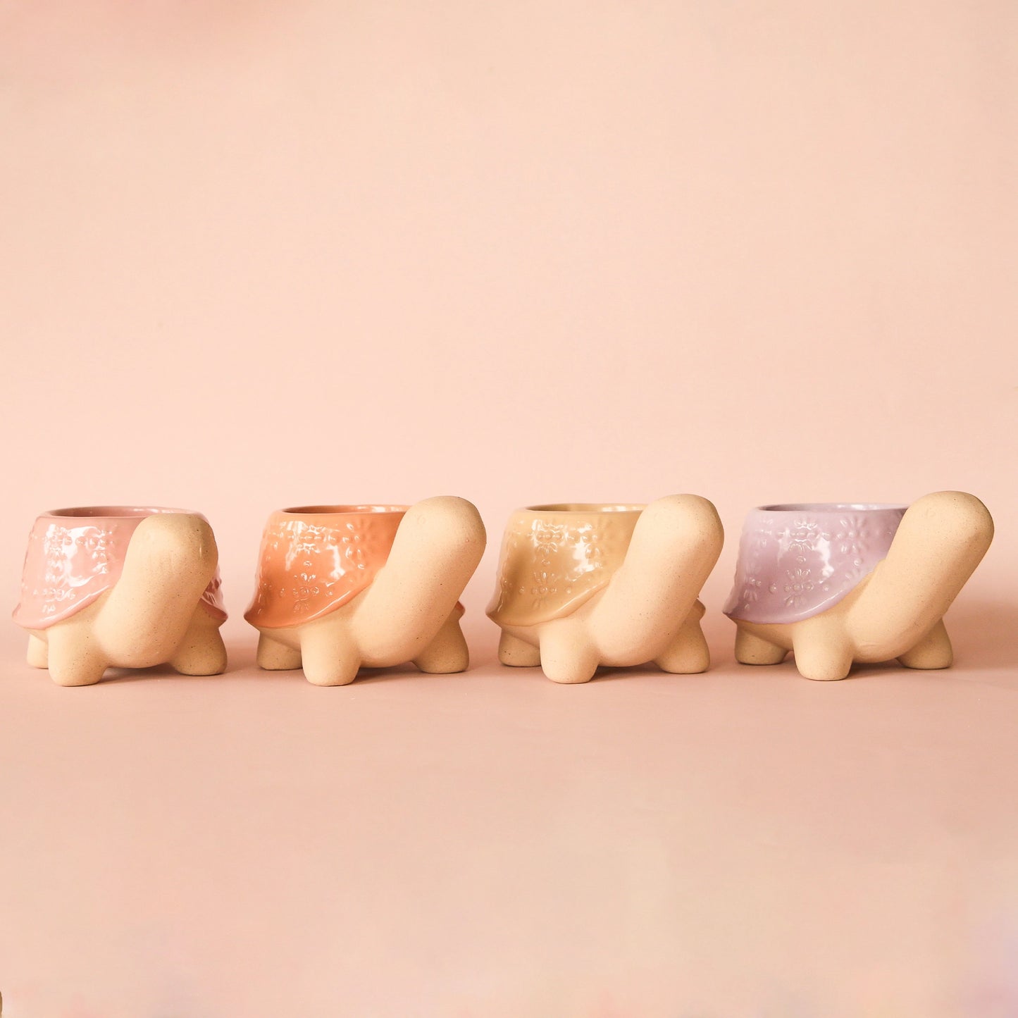On a peachy background is four different colored turtle shaped ceramic planters. From left to right, the colors are pink, sunset, sand and lilac.