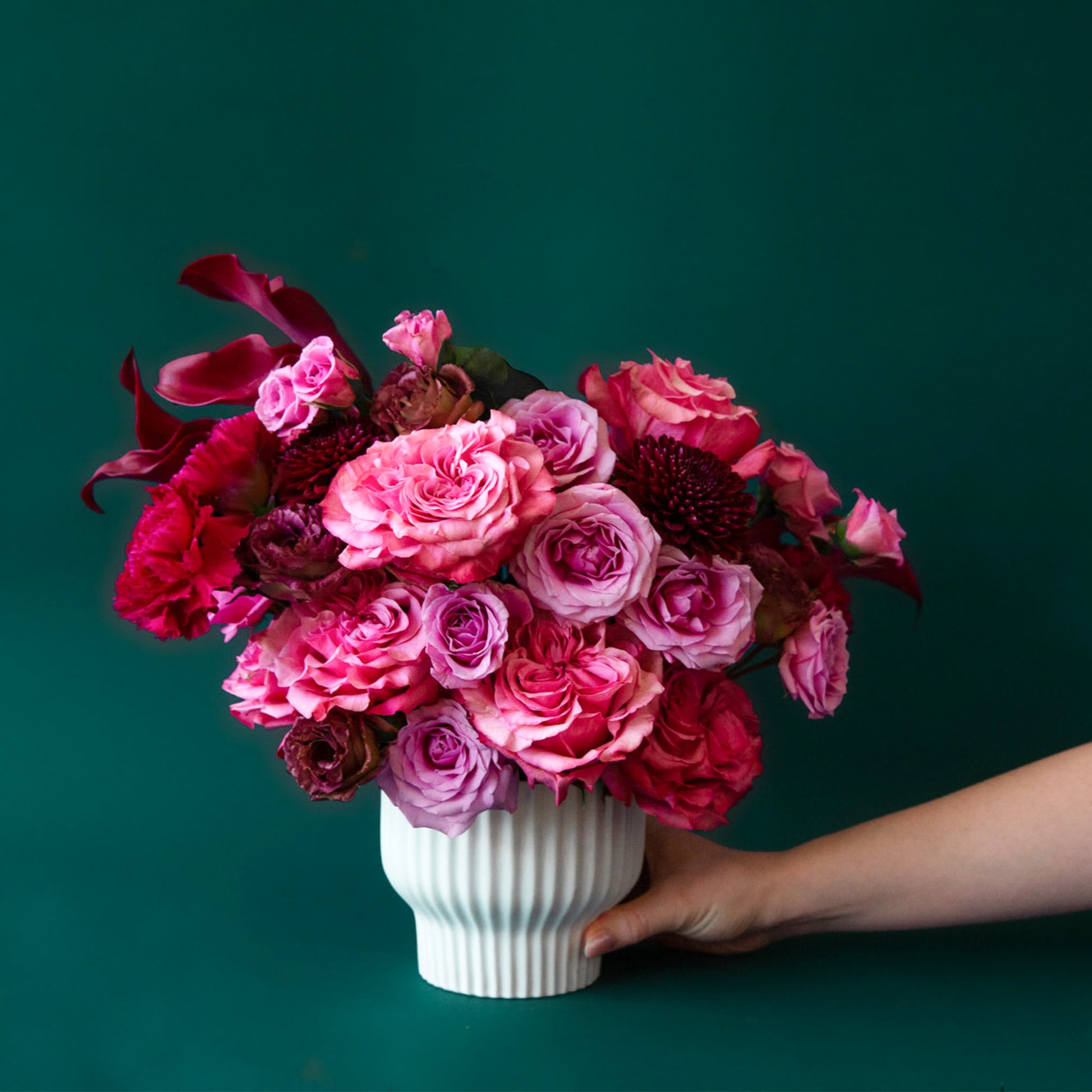 On a blue background is a white ribbed pedestal planter filled with flowers. 