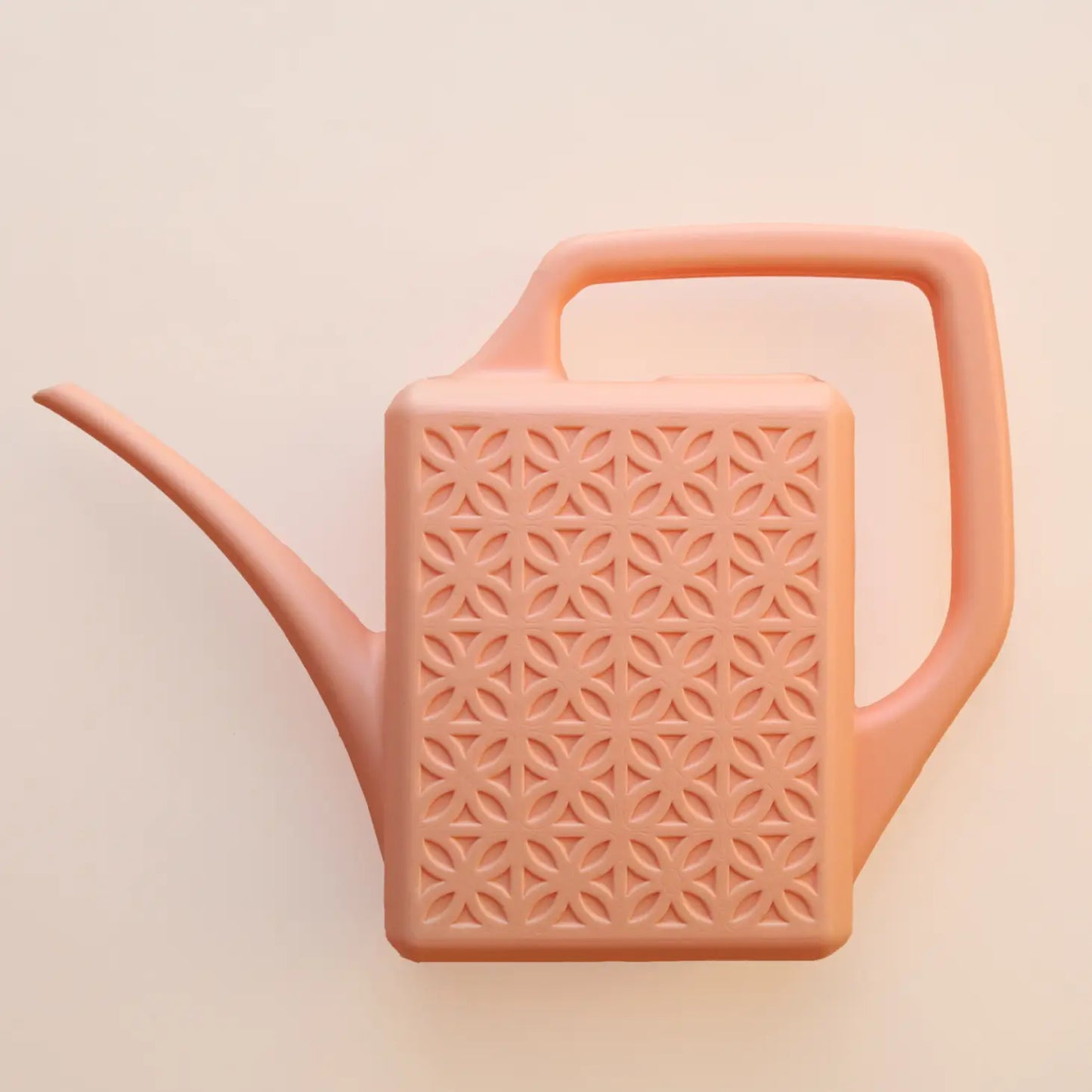On a peachy background is  a light pink plastic watering can with a breeze block design on both sides along with a long spout and a squared off handle for easy watering.
