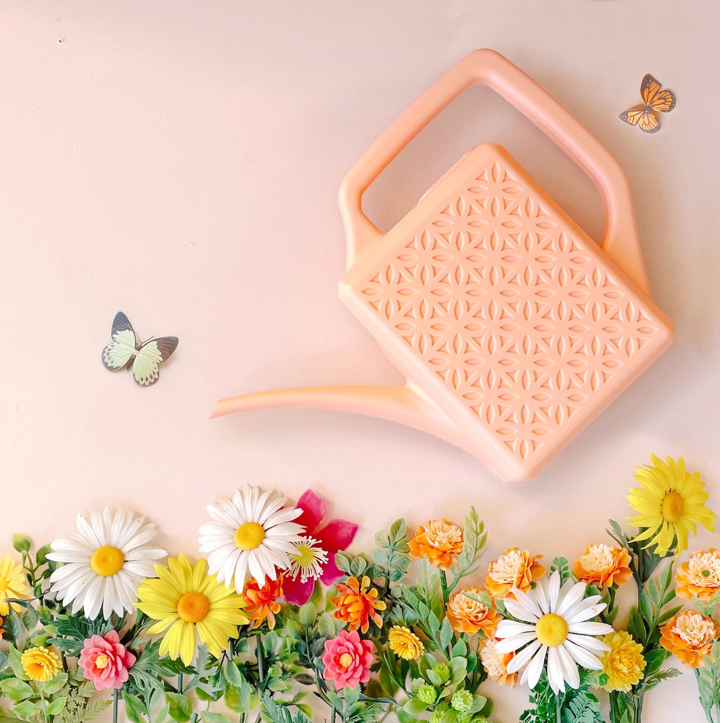On a pink background is a pink plastic watering can with a breeze block design on both sides along with a long spout and a squared off handle for easy watering. It's staged on top of a row of colorful flowers as if its watering alongside two butterflies.