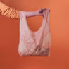 On a orange background is a light pink reusable tote bag made of nylon and detailed with line drawings of alocasia leaves. 