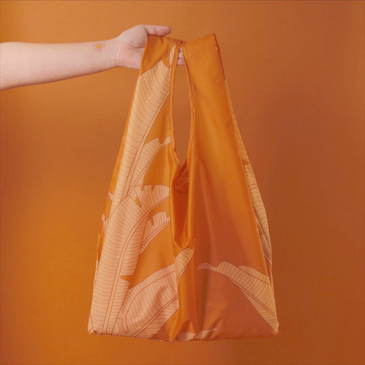 Photographed in front of an orange background is an orange nylon reusable bag with two straps and a cream bird of paradise print on the front and back.