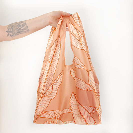 On a white background is a light orange reusable nylon tote bag with line drawings of banana leaves.
