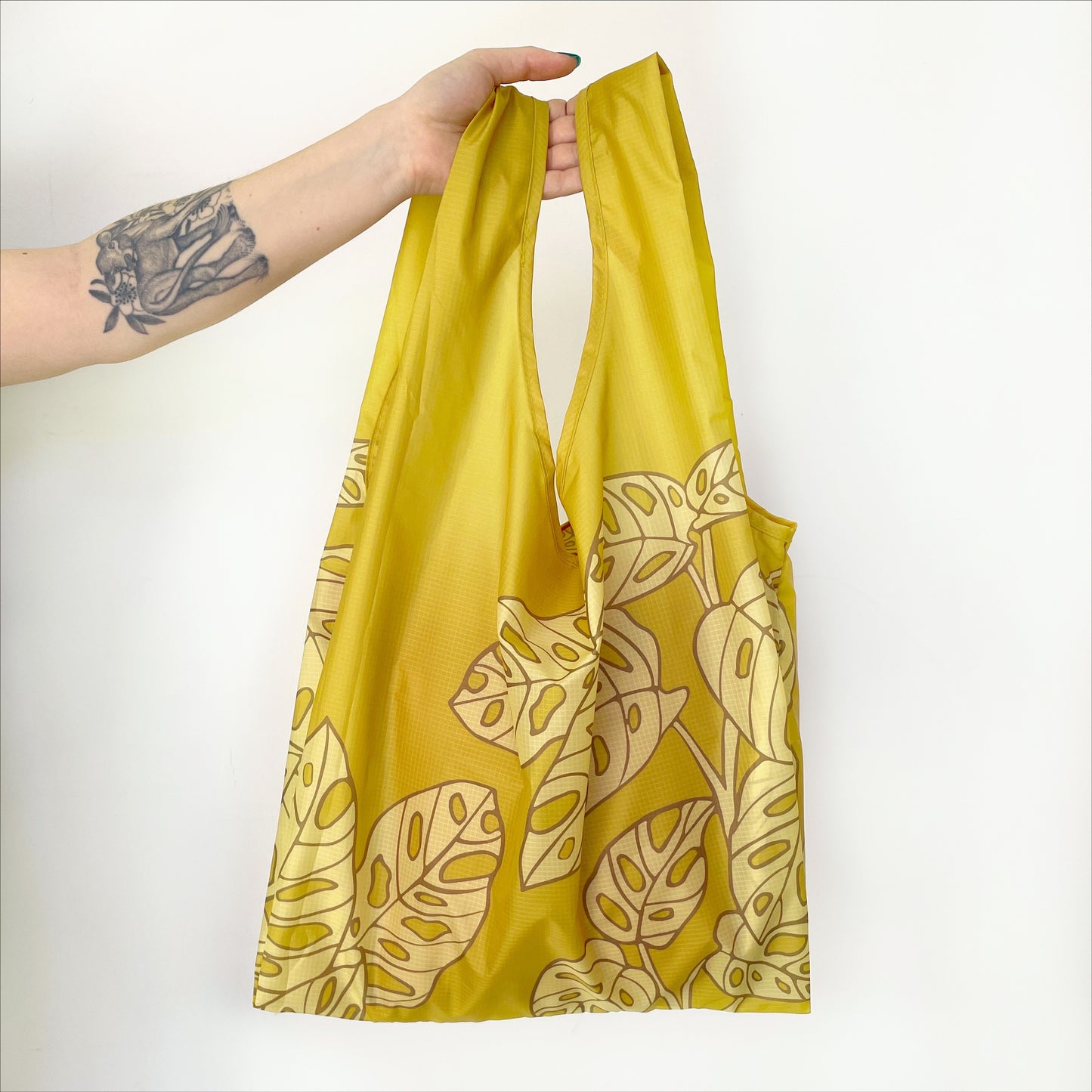 Photographed in front of a white background is a green nylon reusable bag with a lighter green Swiss cheese plant print on the front and back.
