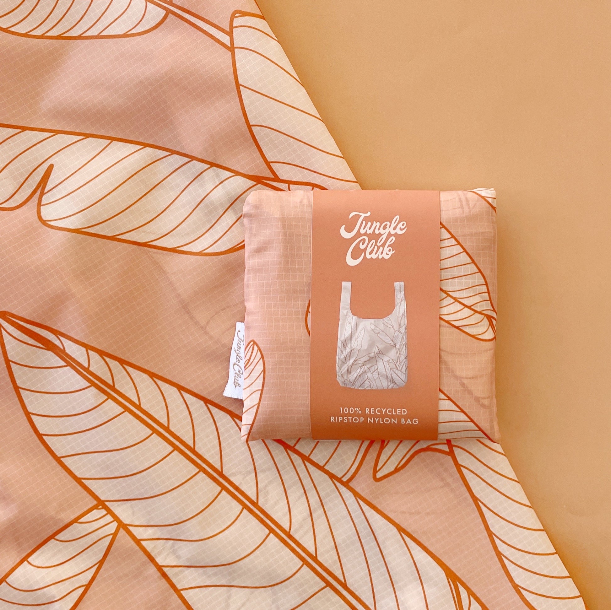 A light orange reusable nylon tote bag with line drawings of banana leaves also folded up and in the condensed foldable pouch.
