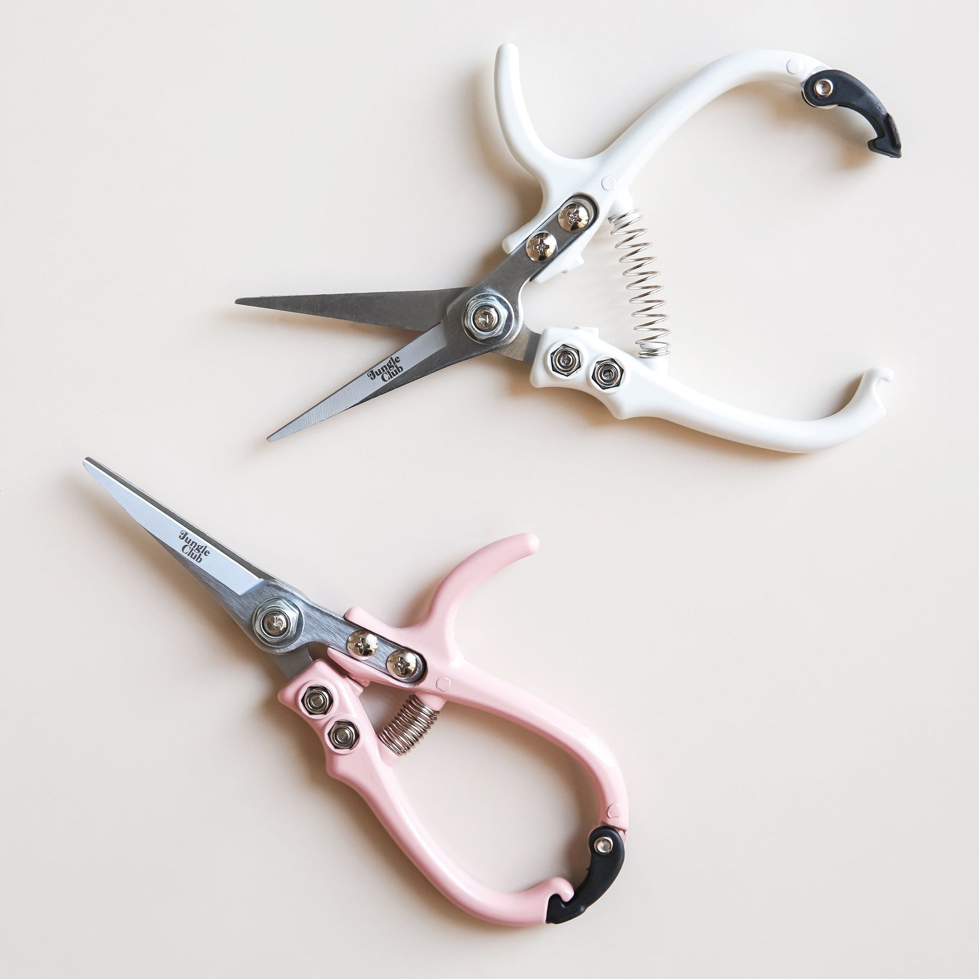 On a cream background is a white pair of pruning shears with silver metal and fixtures and small text on the metal part of the shears that reads, "Jungle Club" in small black letters. In this picture they are photographed next to the same shears in a light pink shade.