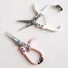On a cream background is a white pair of pruning shears with silver metal and fixtures and small text on the metal part of the shears that reads, "Jungle Club" in small black letters. In this picture they are photographed next to the same shears in a light pink shade. 