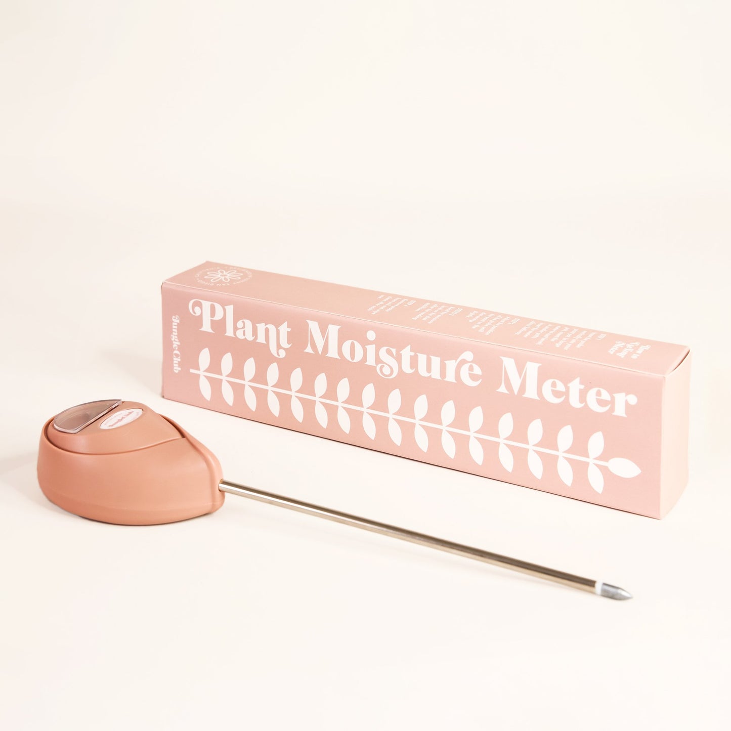 Soft pink moisture meter laying besides packaging that reads 'plant moisture meter' along the side of white leafy design. The moisture meter has an upside down teardrop shape and metal probe running from base.