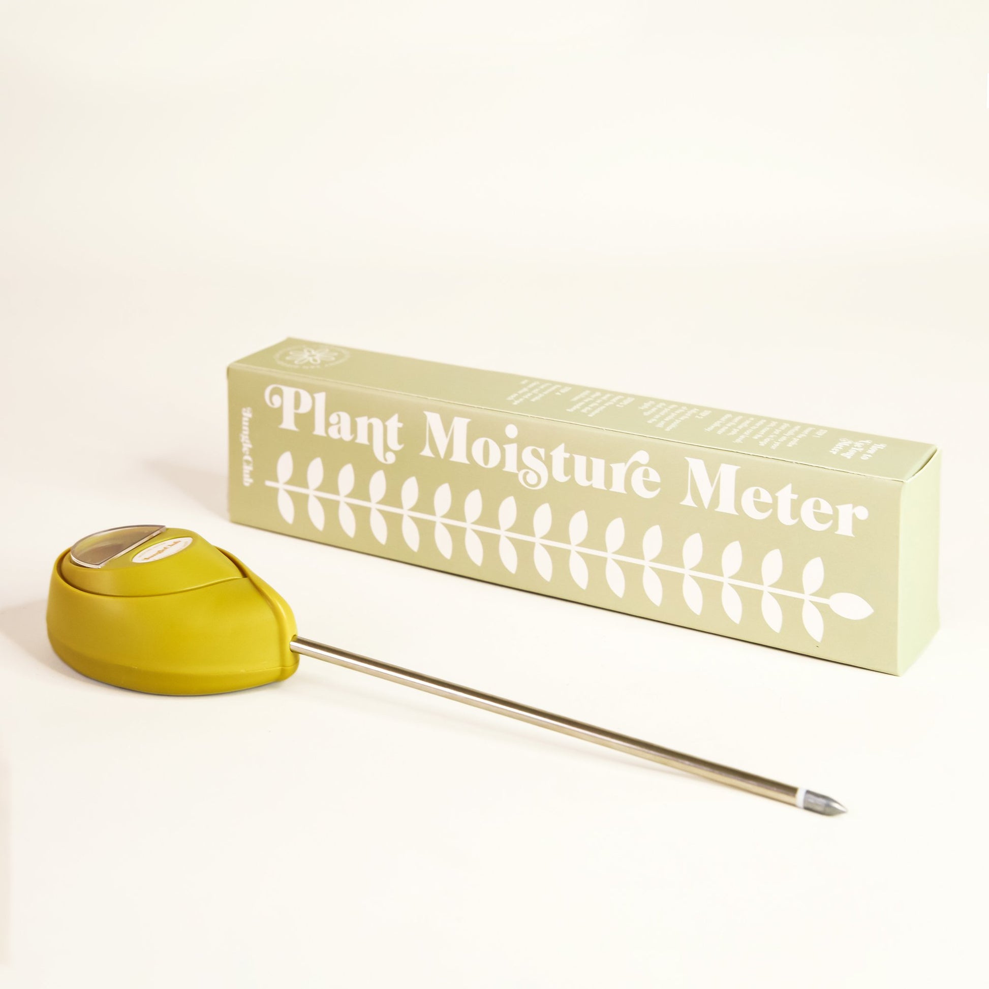 Green moisture meter laying besides packaging that reads 'plant moisture meter' along the side of white leafy design. The moisture meter has an upside down teardrop shape and metal probe running from base. 