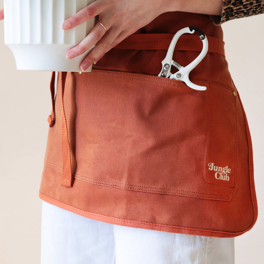 Rust colored gardening apron with 4 pockets. The ties are able to be wrapped around your waist if you would like for a cute bow in the front. There are white pruning shears in the pocket, showing its depth.